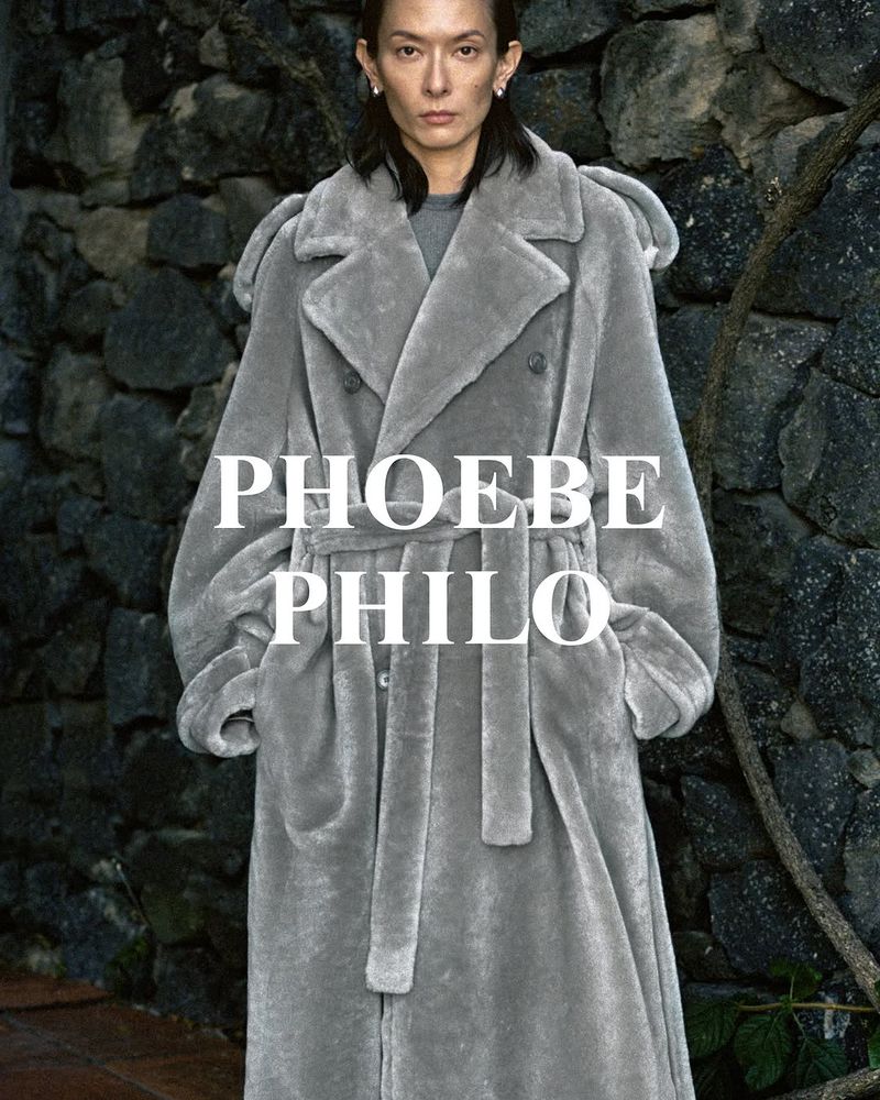 Woman in a grey coat stands against a stone wall. The text "PHOEBE PHILO" is displayed across the center of the image.