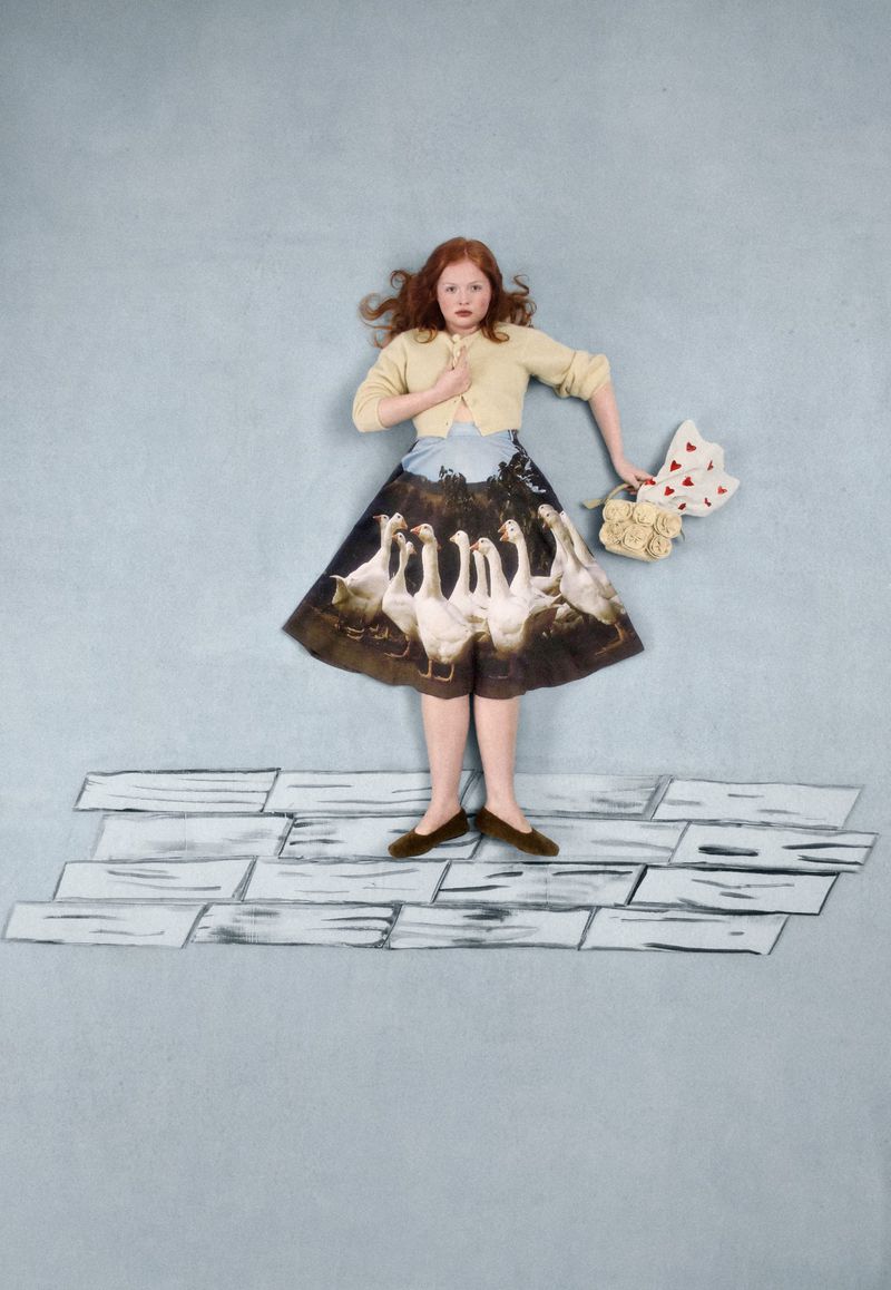 Red-haired woman stands on painted brick floor in pale yellow cardigan, skirt with swan graphic, bouquet and heart-shaped bag.