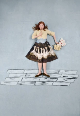 Red-haired woman stands on painted brick floor in pale yellow cardigan, skirt with swan graphic, bouquet and heart-shaped bag.