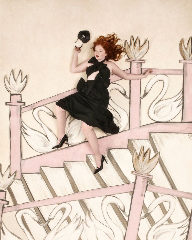Woman in black dress lies on a swan-themed staircase holding a baseball cap in a fashion editorial setting with surreal elements.