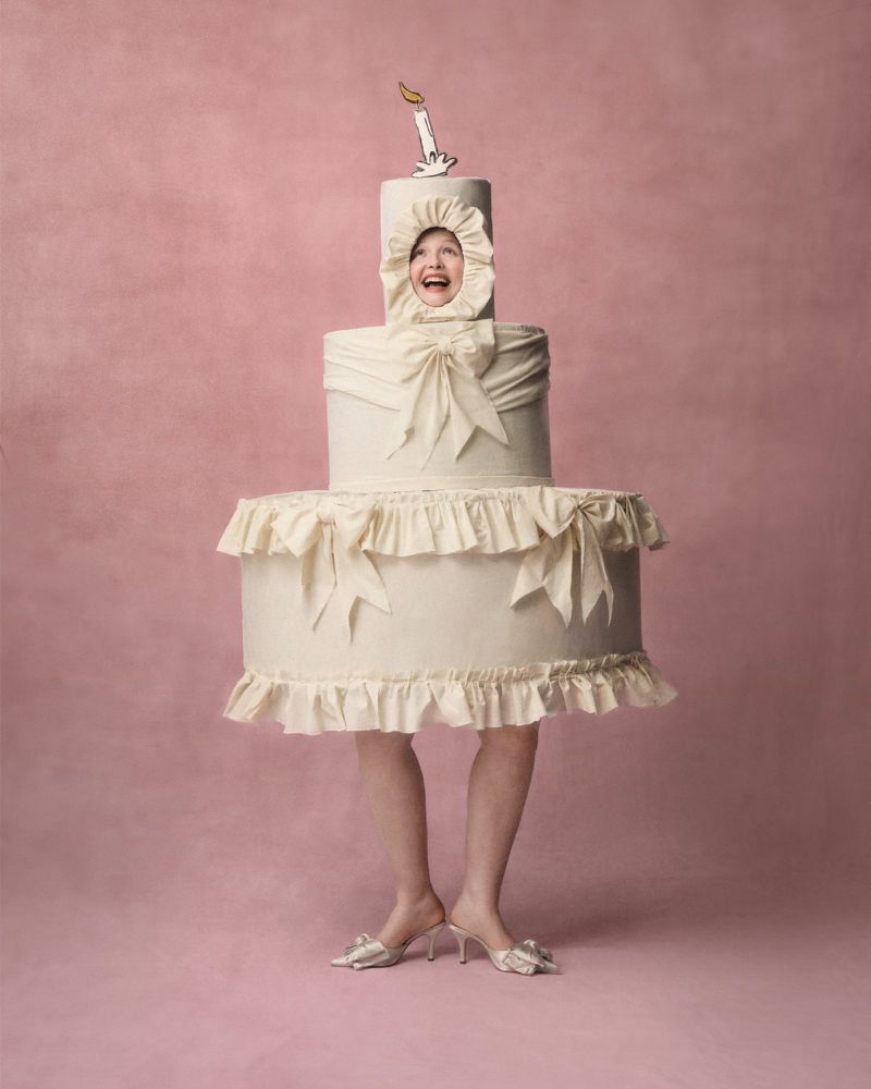 A woman with a big smile wears a cake costume on a pink backdrop, creating a whimsical and fun portrait with studio lighting.