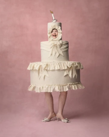 A woman with a big smile wears a cake costume on a pink backdrop, creating a whimsical and fun portrait with studio lighting.