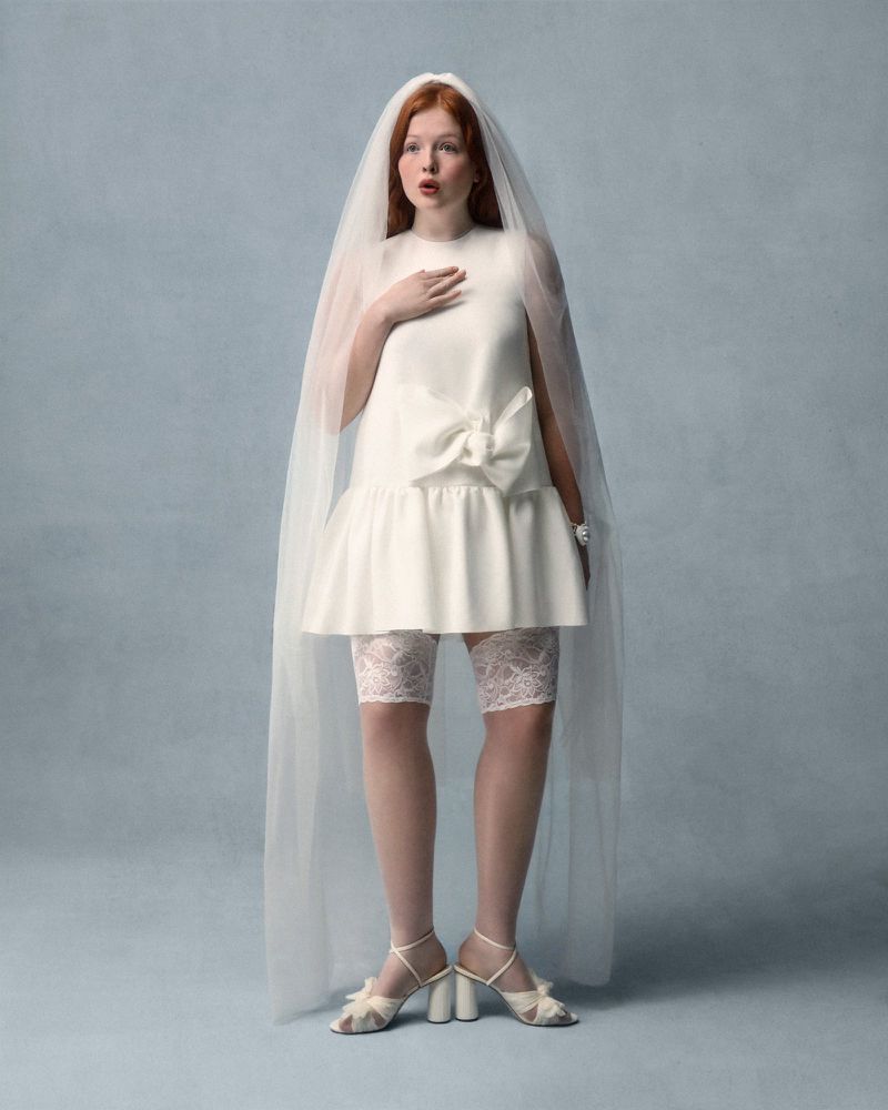 Full-length portrait of a young woman in a short white wedding dress and veil, standing in a studio setting with a light blue background.