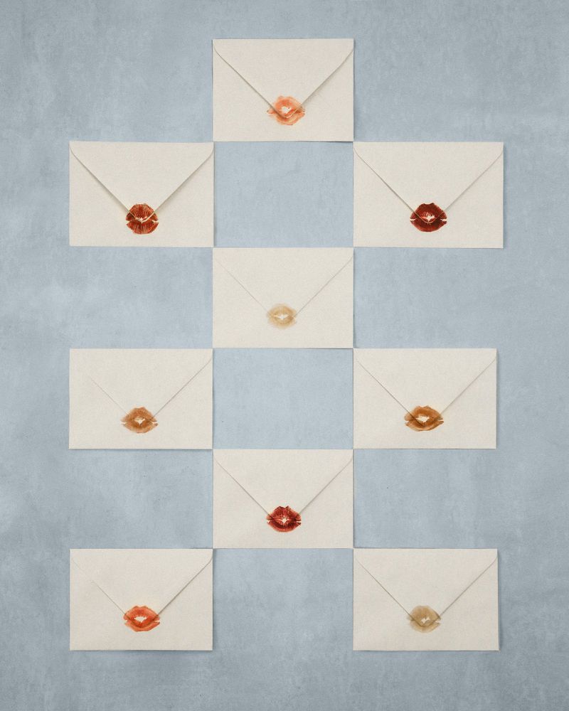 Arrangement of sealed envelopes with lipstick marks in varying shades, symmetrically patterned against a grey textured background.