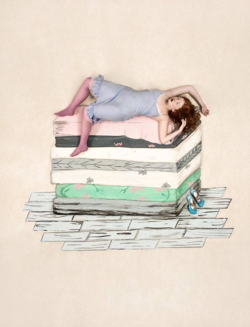 A woman with red hair lies atop a stack of mattresses, wearing a vintage-style negligee and stockings, with shoes on a painted floor.