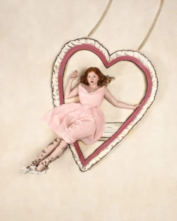 A red-haired young woman in a pink dress poses within a large heart-shaped frame suspended by rope against a neutral backdrop.