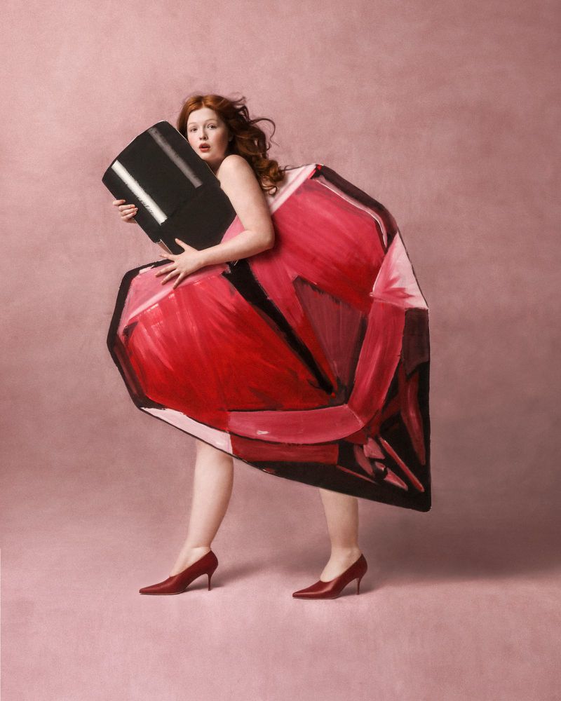 Woman in red heels holding an oversized red nail polish bottle against a light pink background in a beauty or fashion shot.