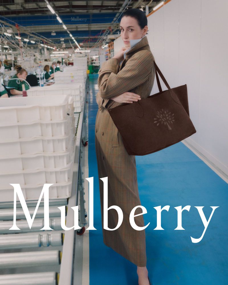 Fashion editorial shot of a woman in a houndstooth coat holding a brown leather Mulberry tote bag in a factory setting.