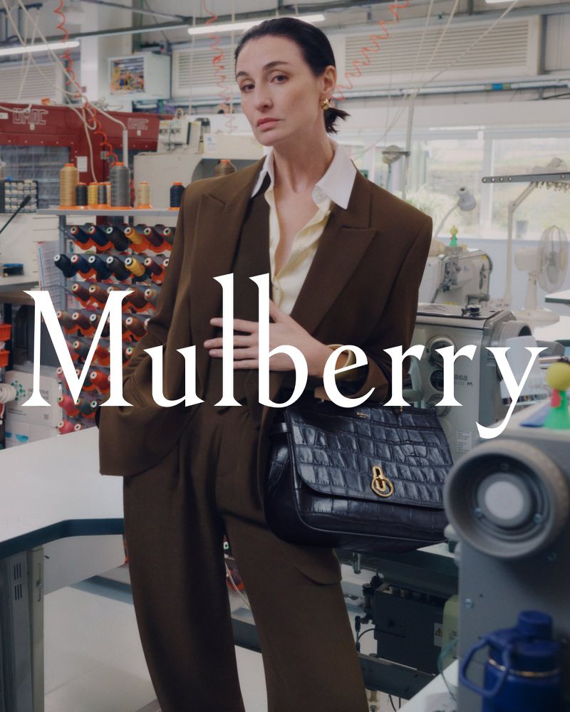 A woman in a brown pantsuit poses indoors holding a black leather handbag with a gold lock detail, in front of sewing equipment.