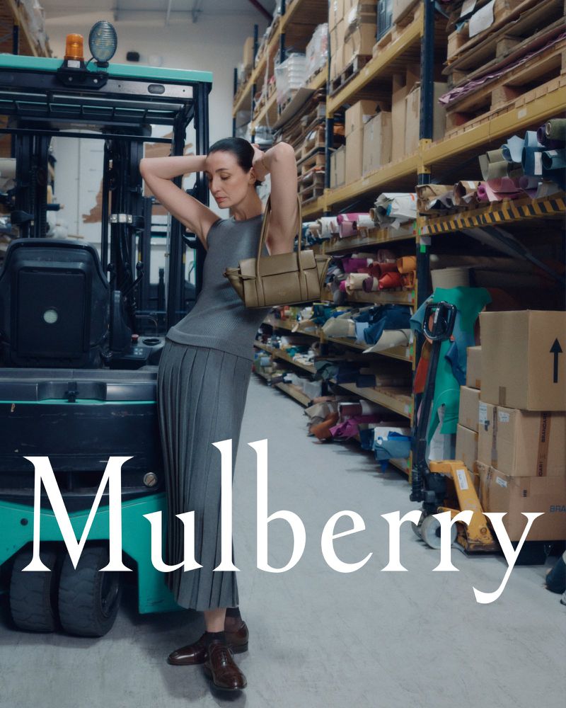A woman models a grey pleated skirt and top with a tan handbag, standing near a teal forklift in a warehouse filled with products.