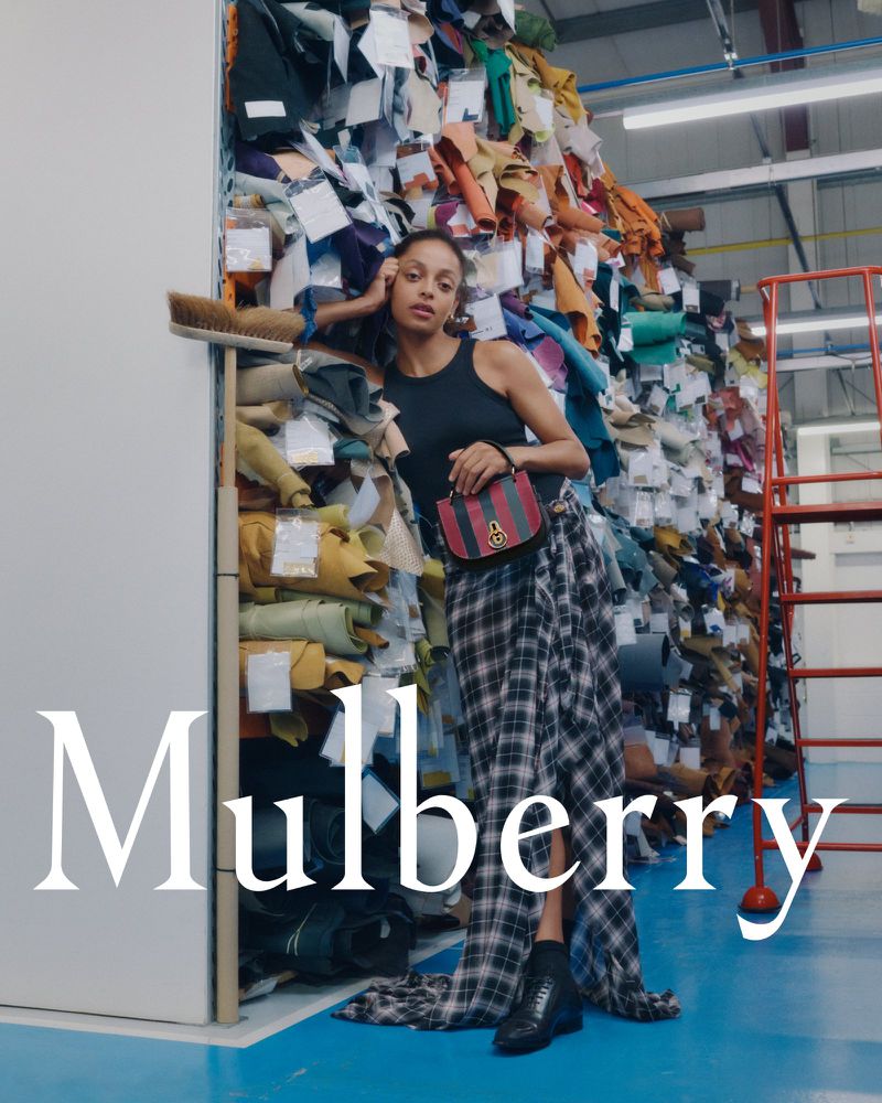 Fashion shot of a Black woman wearing a tank top, plaid skirt, and boots, posing against a backdrop of stacked textiles and materials.