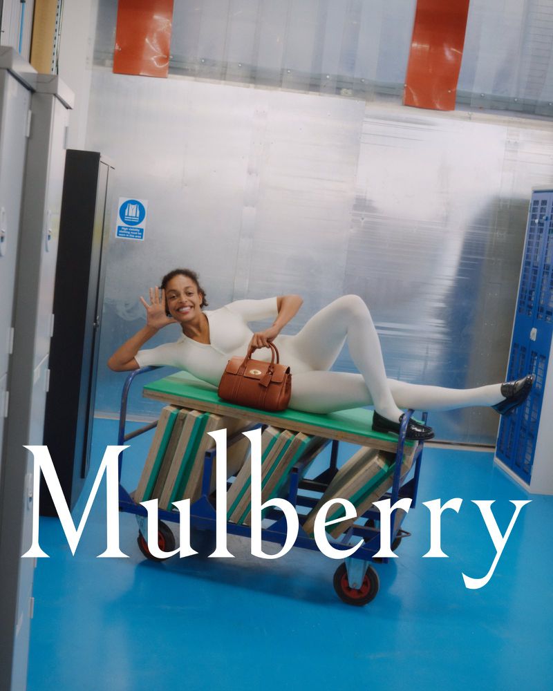 A woman in a white jumpsuit smiles while lying on a blue cart in a studio with a brown leather purse and the word Mulberry.