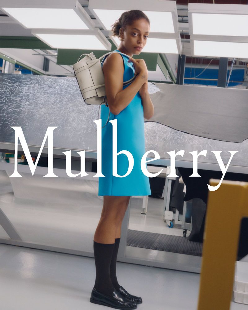 Full shot of an adult female fashion model wearing a teal dress with a white purse in an indoor studio, with 'Mulberry' text overlay.