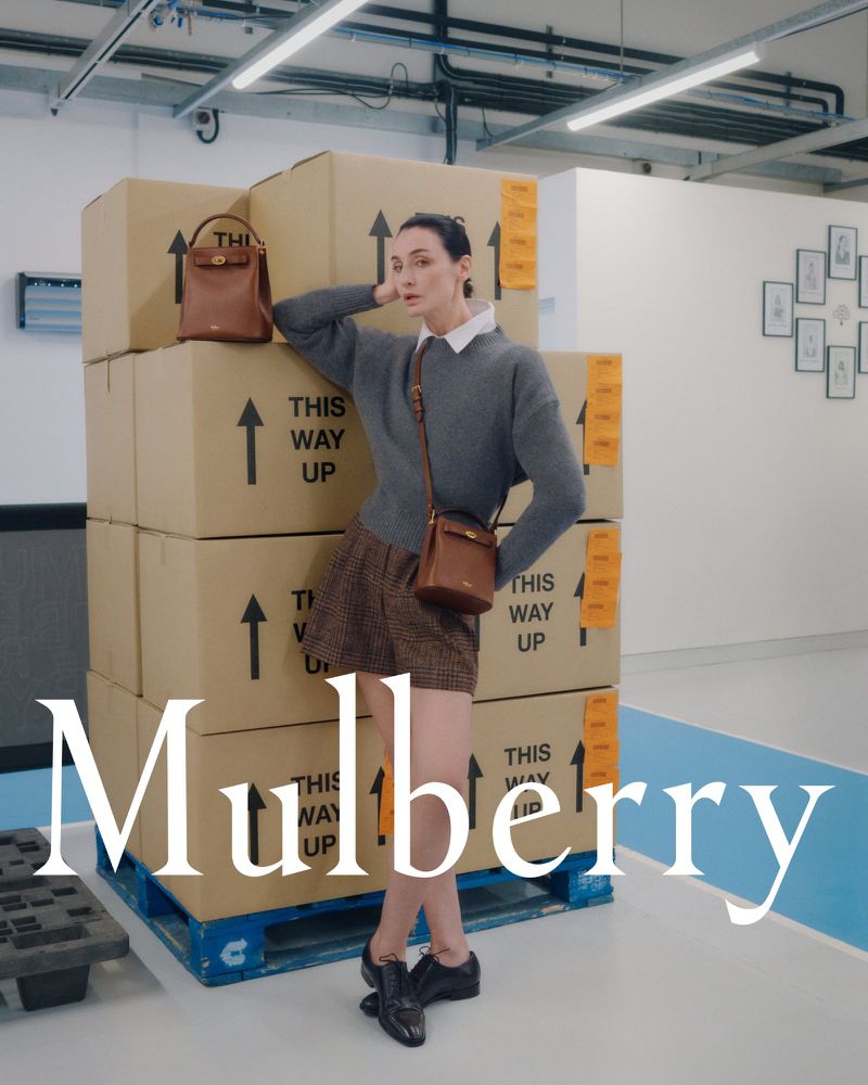 Fashion shot of a woman in brown shorts and a grey sweater leaning against a stack of cardboard boxes with a brown leather purse.