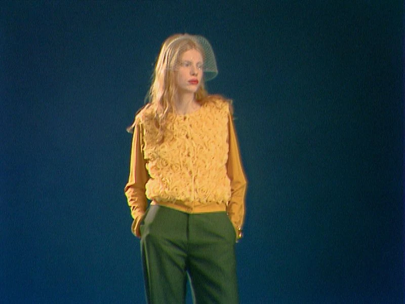 A fair-skinned woman with long blonde hair in a yellow floral vest and green pants posing against a dark teal backdrop.