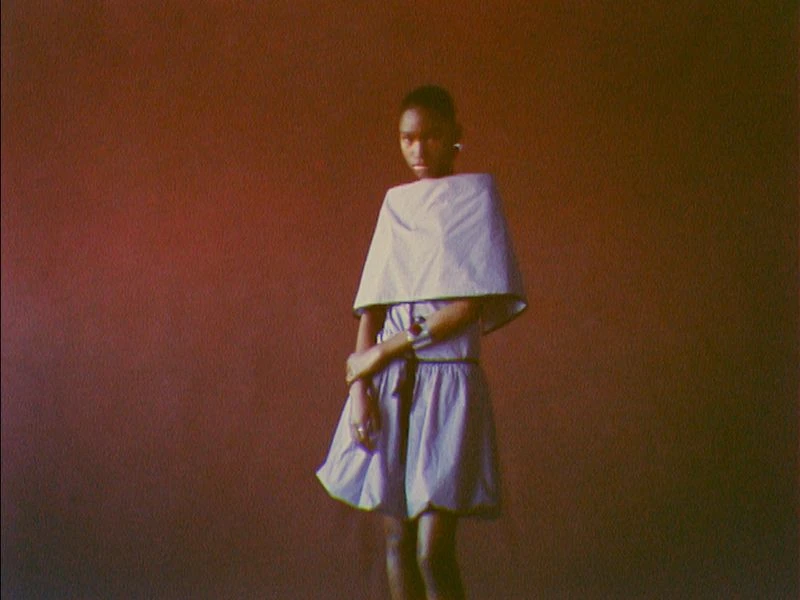 Young woman in a light blue dress with a cape-like collar stands against a warm-toned background, hands crossed, and looks directly at the camera.
