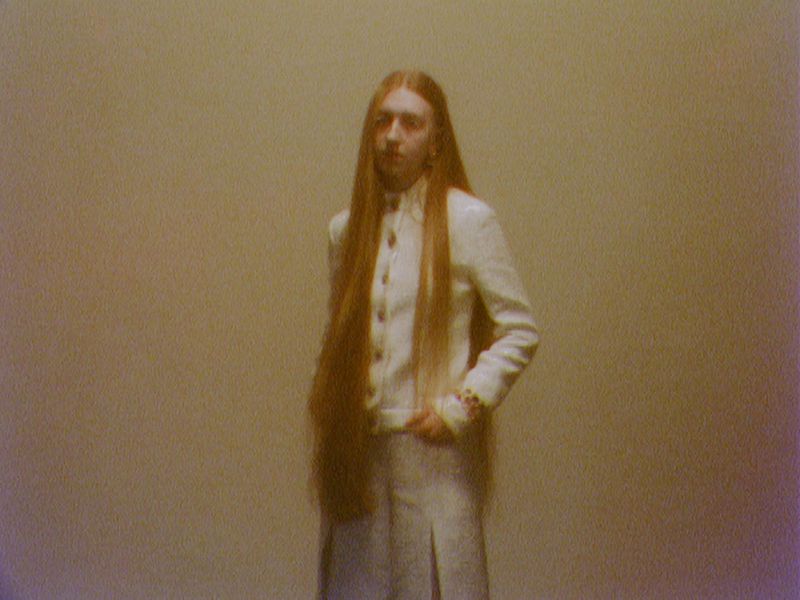 Portrait of a person with long ginger hair, wearing a white suit, standing against a pale background in a softly lit indoor space.