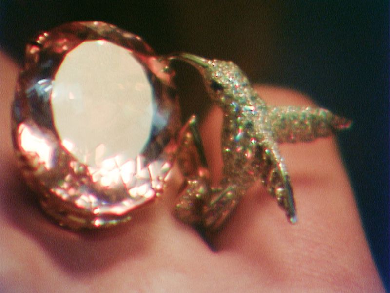 Close-up of a gold ring featuring a large gemstone and a hummingbird design, sparkling and catching the light against skin.