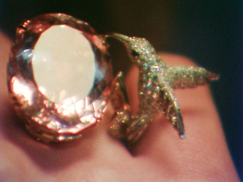 Close-up of a gold ring featuring a large gemstone and a hummingbird design, sparkling and catching the light against skin.