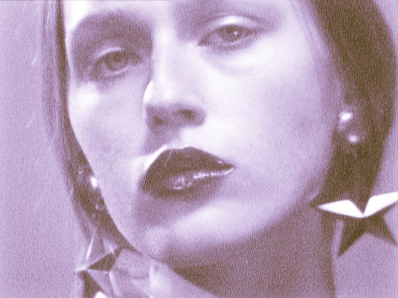 Close-up portrait of a woman with glossy lips, star earrings, and lilac-toned skin, showcasing fashion and beauty elements.