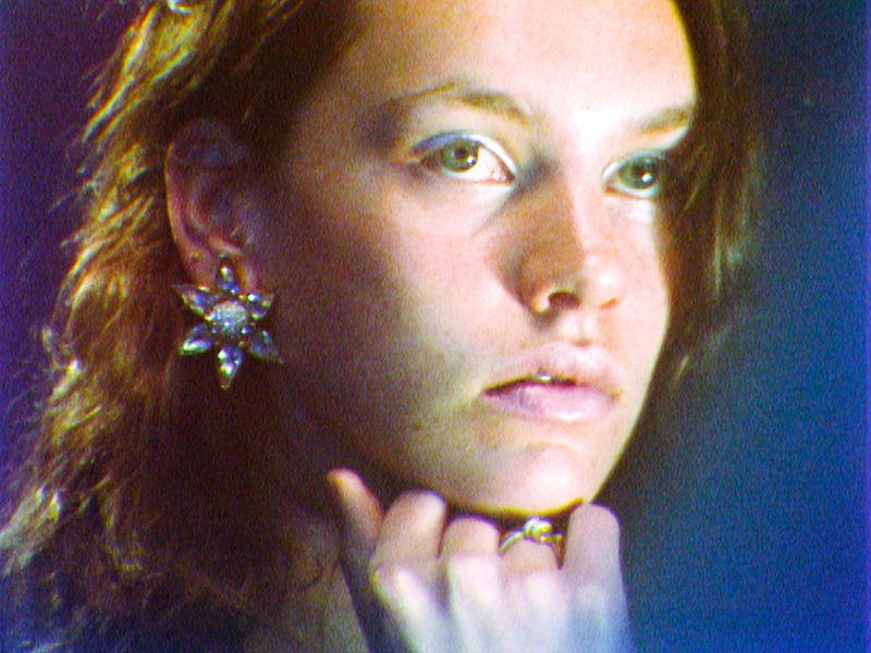 Close-up portrait of a fair-skinned adult female with light brown hair and light eye makeup, wearing flower earrings and a ring.