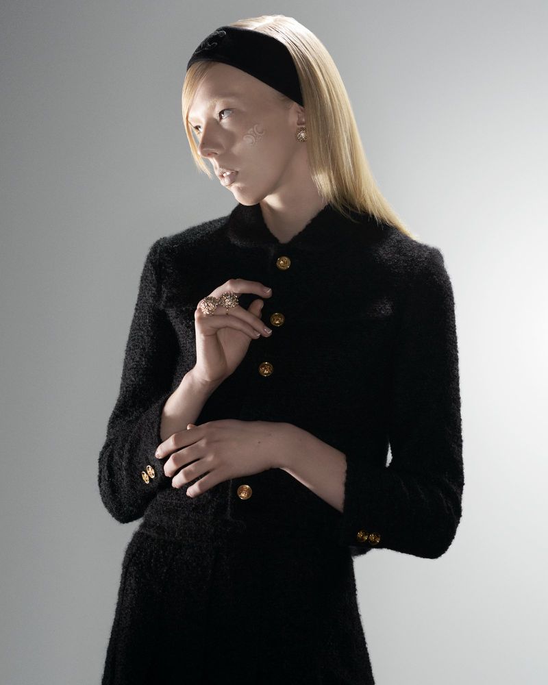 Portrait of a young woman with long blonde hair wearing a black headband and tailored suit jacket with gold buttons in a studio setting.