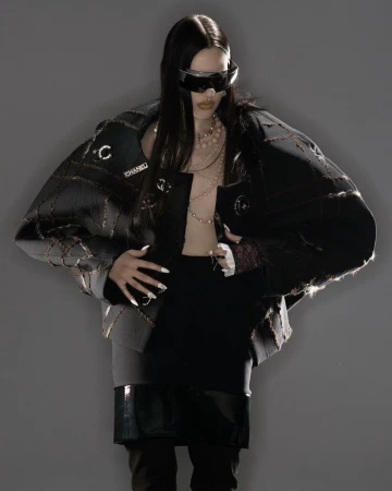 Woman in oversized black jacket, black top, skirt, futuristic glasses, posing against gray backdrop with layered necklaces and white nails.
