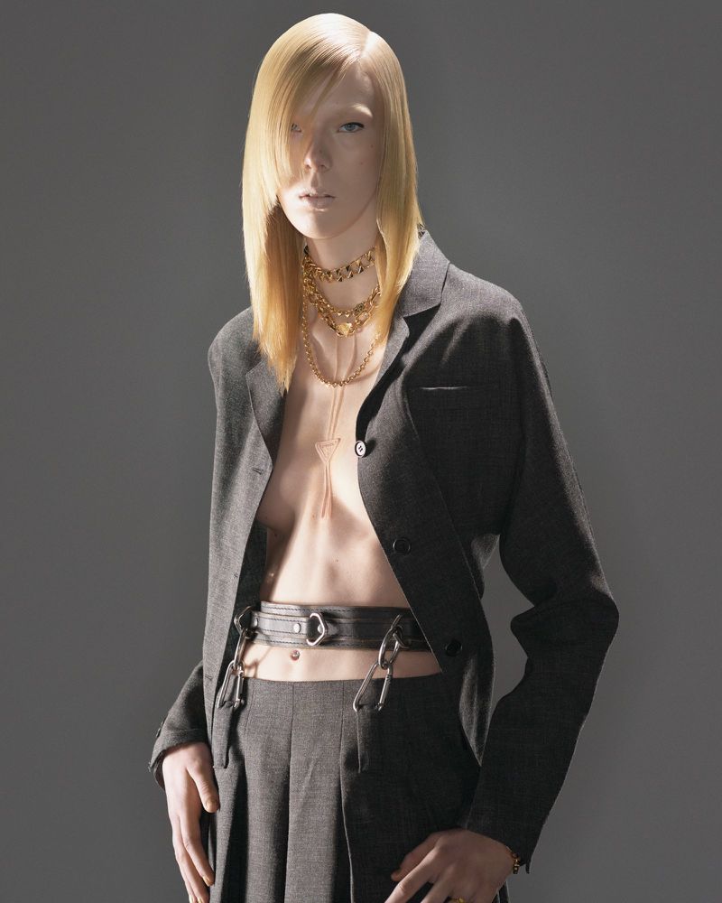 Fashion portrait of a slender model with platinum blonde hair wearing a dark grey blazer and trousers with a belt and layered necklaces.