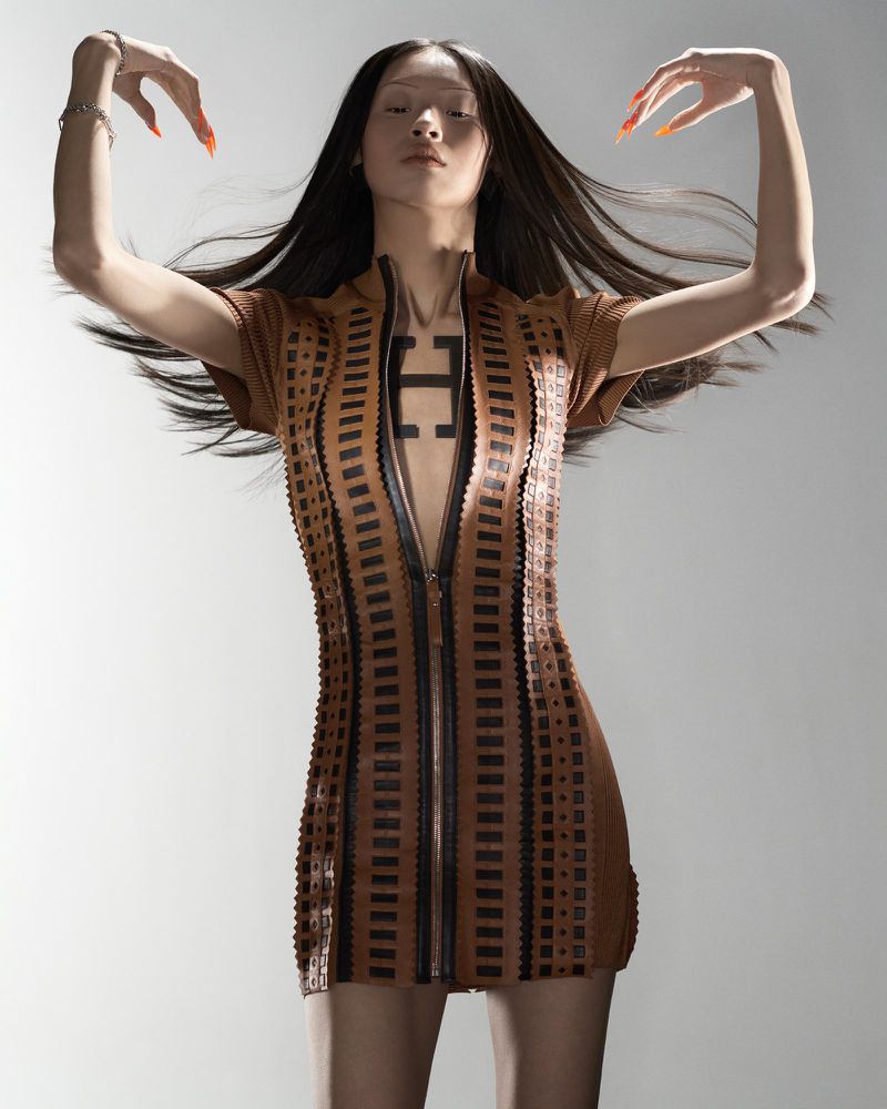 Woman in short brown patterned dress poses with arms raised against a plain background, showcasing fashion and artistic expression.