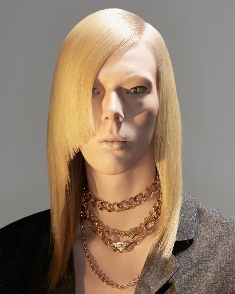 Close-up studio shot of an adult with blond hair styled with a side part, wearing a black jacket and layered gold chain necklaces.