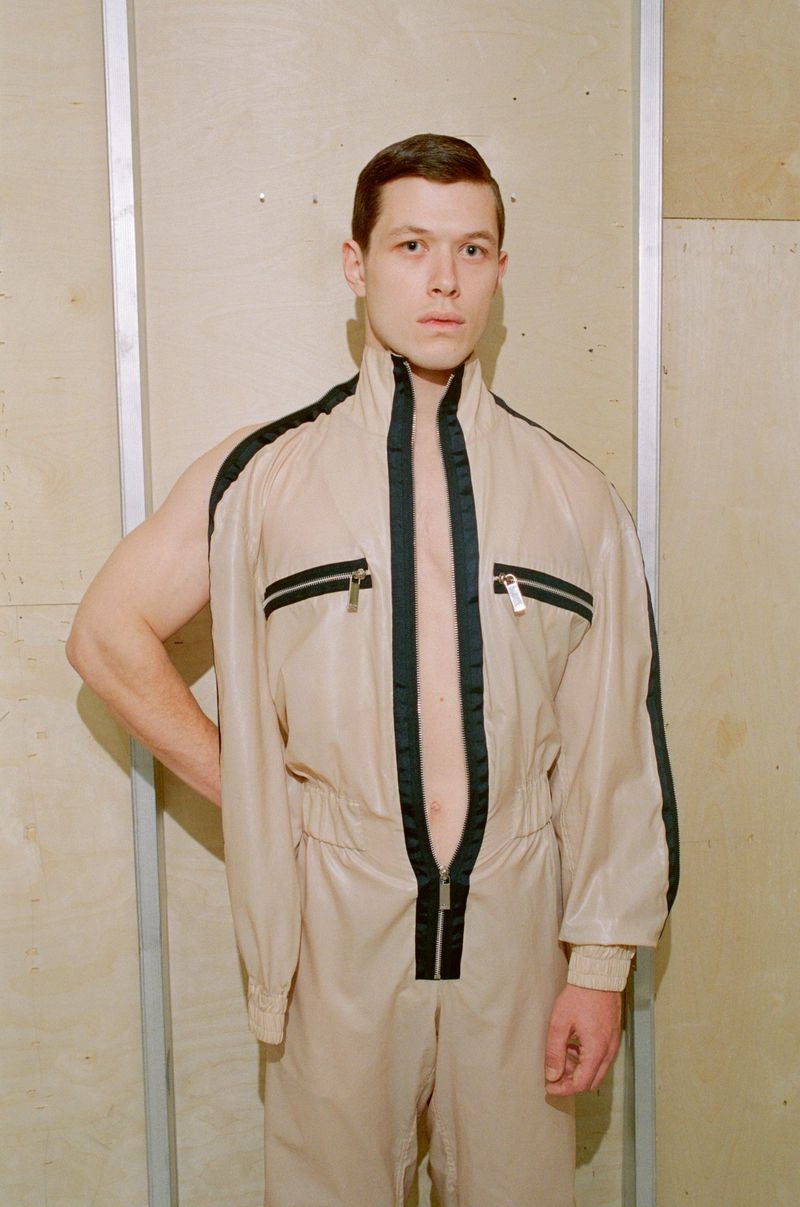 A man in a beige jumpsuit with black stripes stands in front of a wooden wall in a studio setting, posing with one sleeve off.