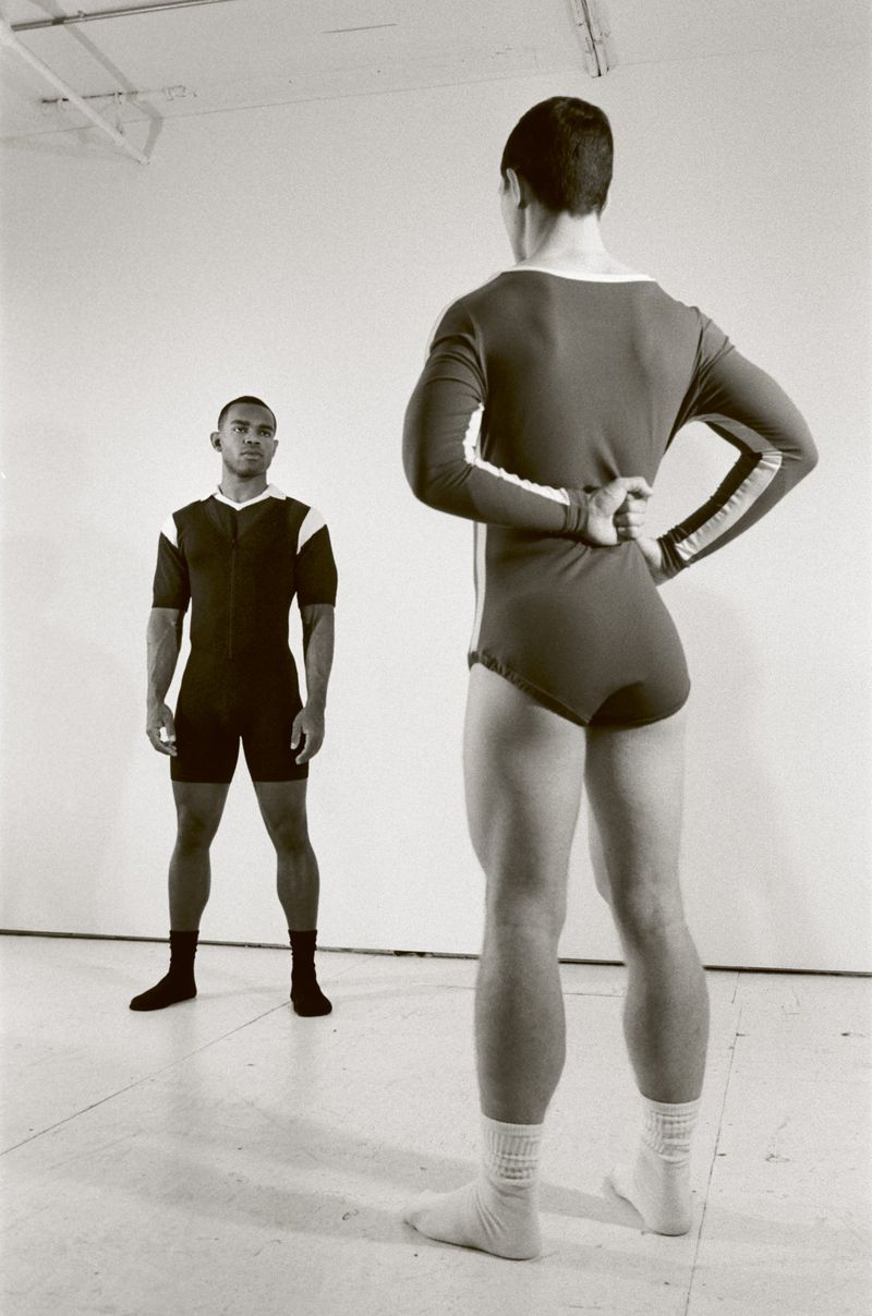 Two men in athletic attire pose in a bright, neutral-toned studio. One faces forward; the other, with hands on his lower back, faces away.