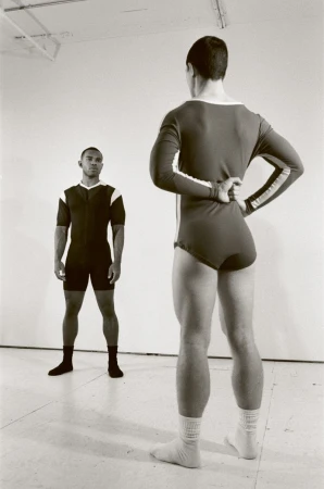 Two men in athletic attire pose in a bright, neutral-toned studio. One faces forward; the other, with hands on his lower back, faces away.