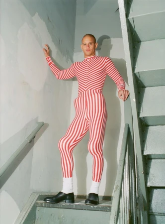 An adult man stands on stairs in a red and white striped bodysuit with black dress shoes, posed indoors with neutral expression.