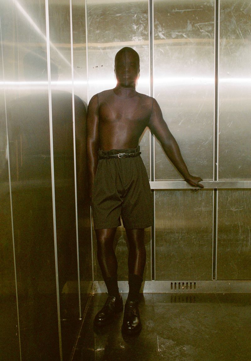 Monochrome portrait of a single adult leaning against a stainless steel elevator wall, wearing shorts, socks, and dress shoes.