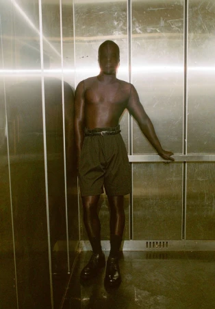 Monochrome portrait of a single adult leaning against a stainless steel elevator wall, wearing shorts, socks, and dress shoes.