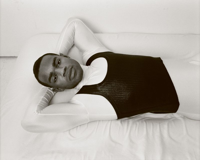 Black and white photograph of a Black adult male in a white long-sleeved shirt and black tank top, reclining on a white surface indoors.