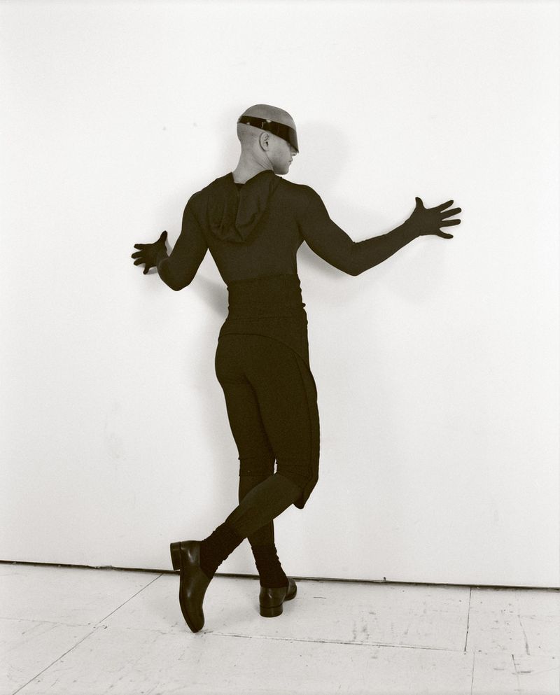 Male in dark body suit poses against white wall, expressive arms, bald head with visor, shiny black shoes and gloves, studio lighting.