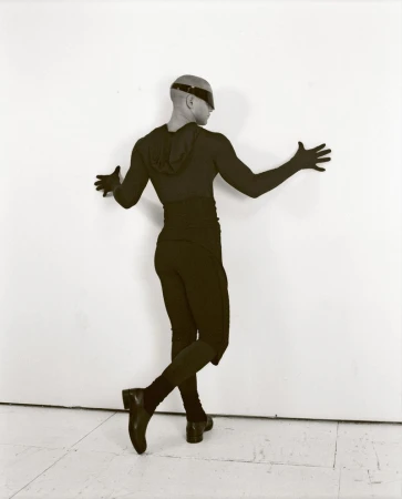 Male in dark body suit poses against white wall, expressive arms, bald head with visor, shiny black shoes and gloves, studio lighting.