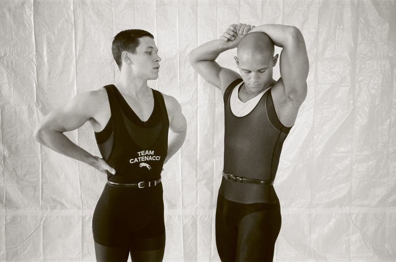Two athletic adult men in black athletic apparel posing in front of white textured backdrop in a brightly lit indoor environment.