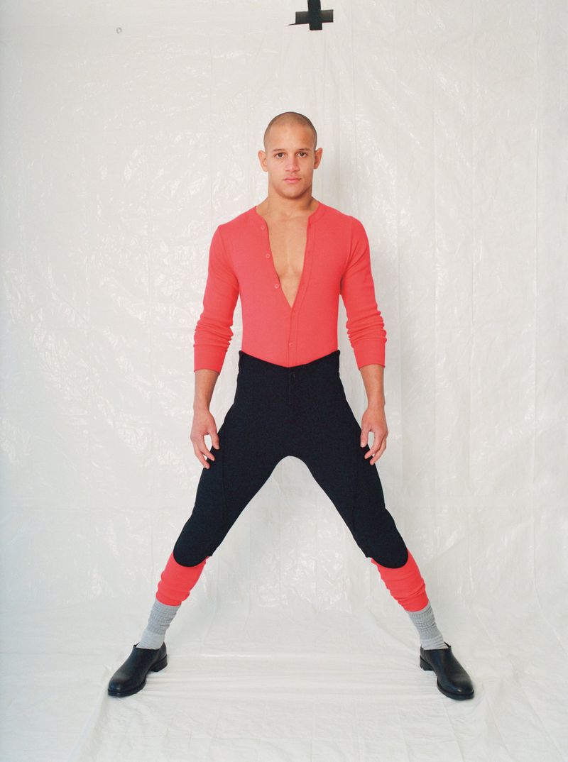 Man in a pink shirt and black pants strikes a wide stance against a white backdrop, creating a visually striking and bold fashion image.