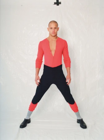 Man in a pink shirt and black pants strikes a wide stance against a white backdrop, creating a visually striking and bold fashion image.
