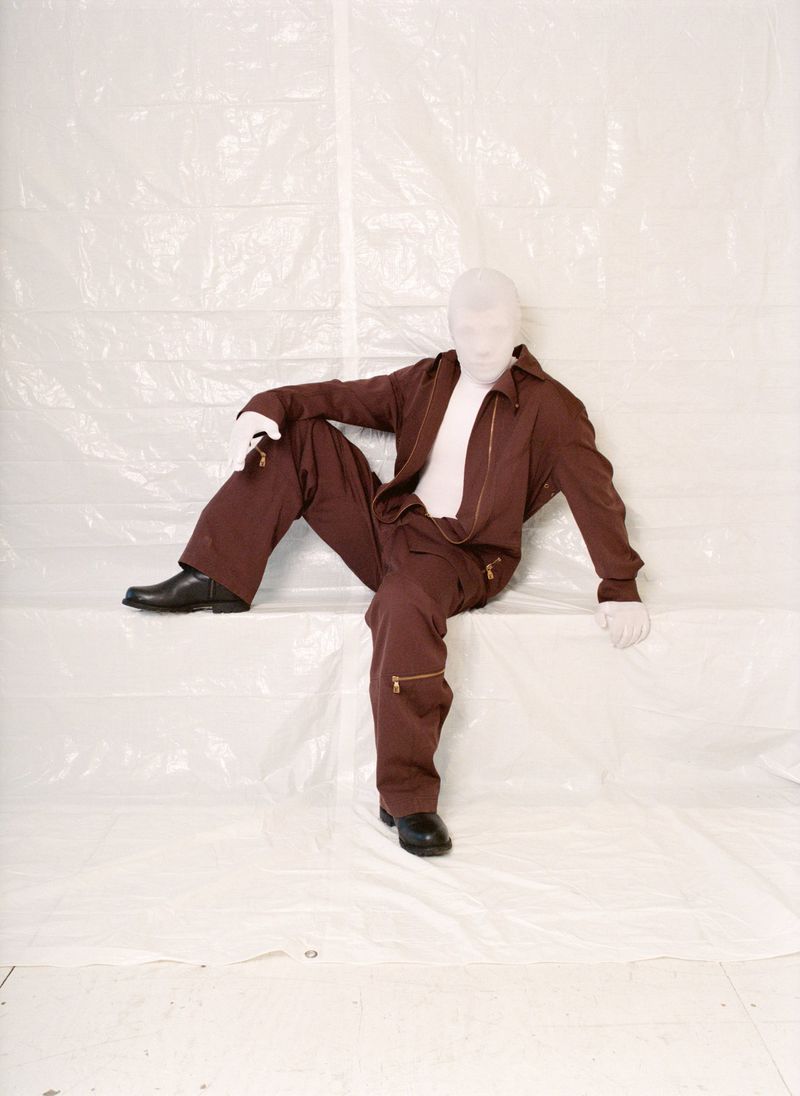 A person in a tan suit and white gloves is seated against a crinkled, white backdrop, with black shoes adding contrast to the neutral tones.