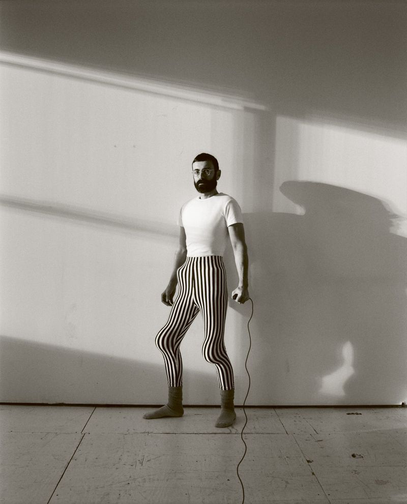 Monochrome portrait of a bearded man in striped leggings and boots, standing in a room with shadows, holding a jump rope.