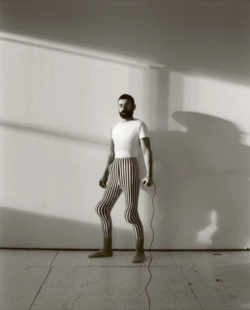 Monochrome portrait of a bearded man in striped leggings and boots, standing in a room with shadows, holding a jump rope.