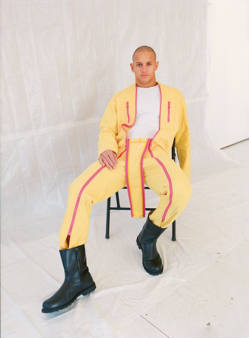 Adult male with shaved head sits in studio, modeling yellow jumpsuit with red trim, white t-shirt, and black boots against white backdrop.