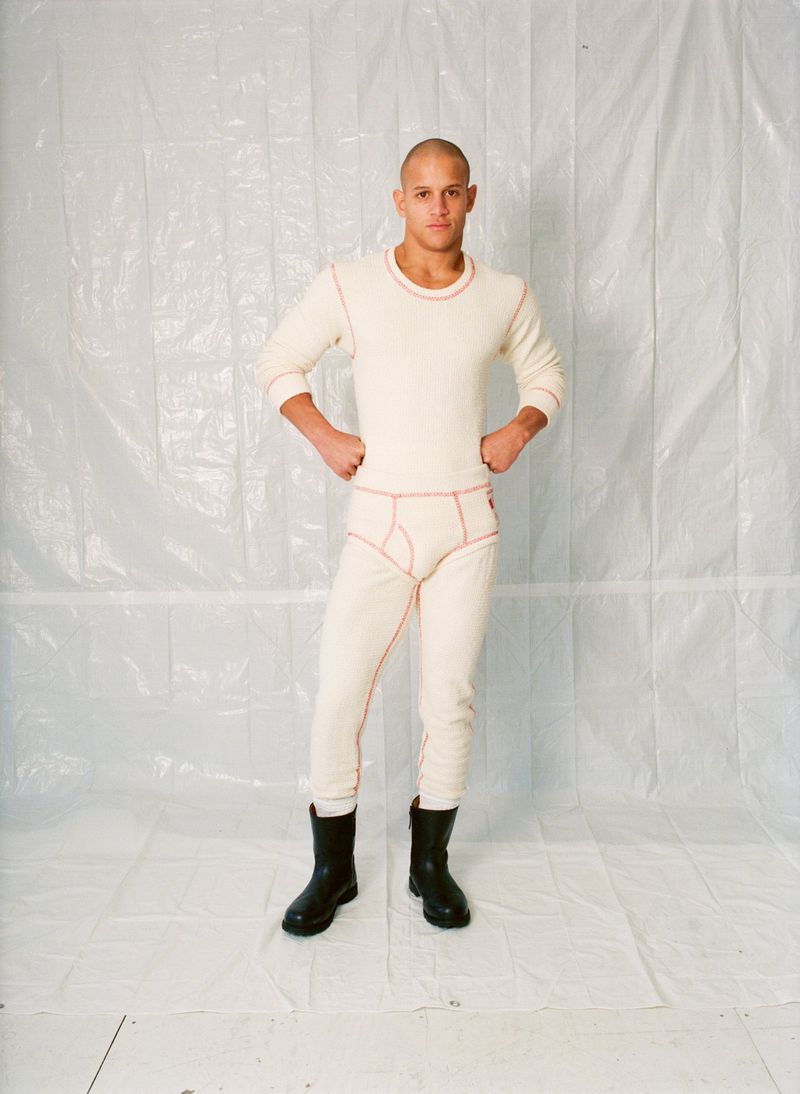Full shot of a male adult in a long sleeve shirt, bottoms, and black boots against a crinkled white background, hands on hips.