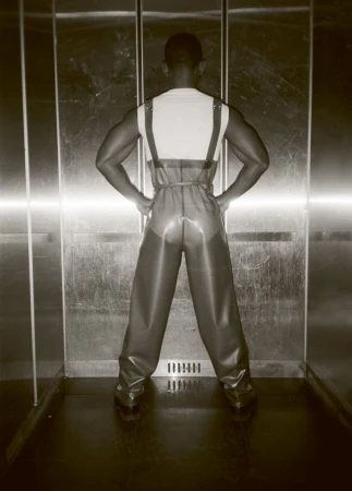 Back view of a muscular adult male in rubber overalls in an elevator, with hands on hips, showcasing a unique fashion statement.