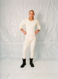 Man wearing cream thermal underwear with red contrast stitching and black boots standing against white plastic background