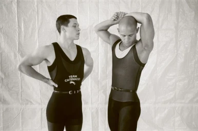 Two men wearing athletic singlets and belts posing against a crinkled white backdrop in a black and white fashion editorial.